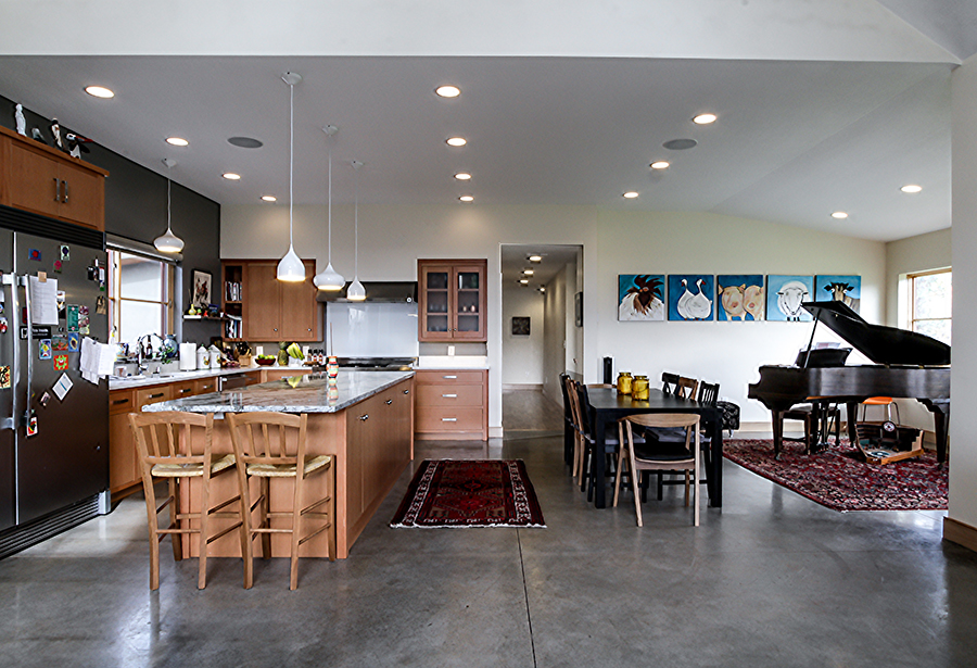 A very open kitchen and dining room with a grey, possibly stone, floor. A kitchen island, stainless steel refridgerator, black dining table, piano, and several decorative rugs are visible.